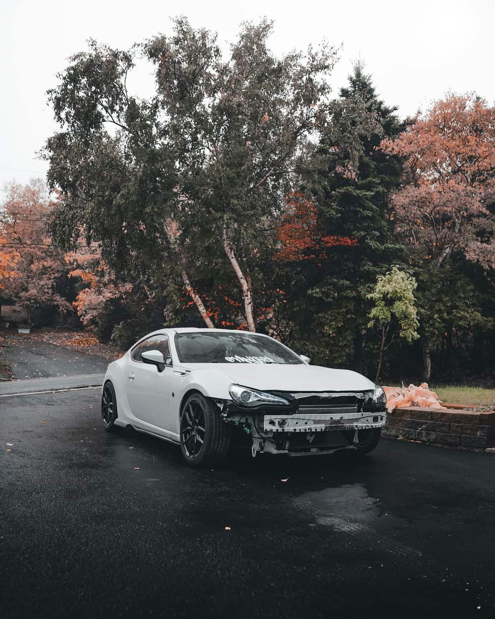 A white car with a damaged front bumper stopped on a wet road on an overcast autumn day.